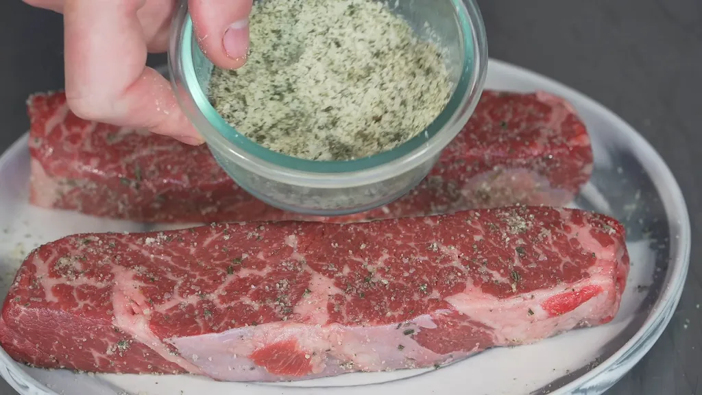 Close-up of raw Denver steaks on a plate being seasoned with a herb-infused salt mixture