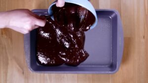 A Person Pours Thick, Glossy Nutella Brownie Batter from A Blue Mixing Bowl Into a Greased Baking Pan