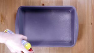 A Hand Sprays Non-Stick Cooking Spray onto A Rectangular Baking Pan on A Wooden Surface