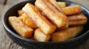 Close-up of perfectly fried yuca fries in a dark ceramic bowl on a rustic wooden surface