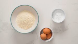 A Bowl of Shredded Coconut, a Small Bowl of Sugar, and A Dish with Three Brown Eggs Are Arranged on A White Countertop