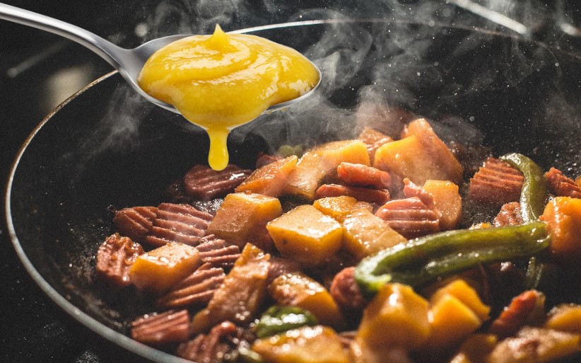 A yellow miso butter is being poured over a medley of cooked vegetables and sausage