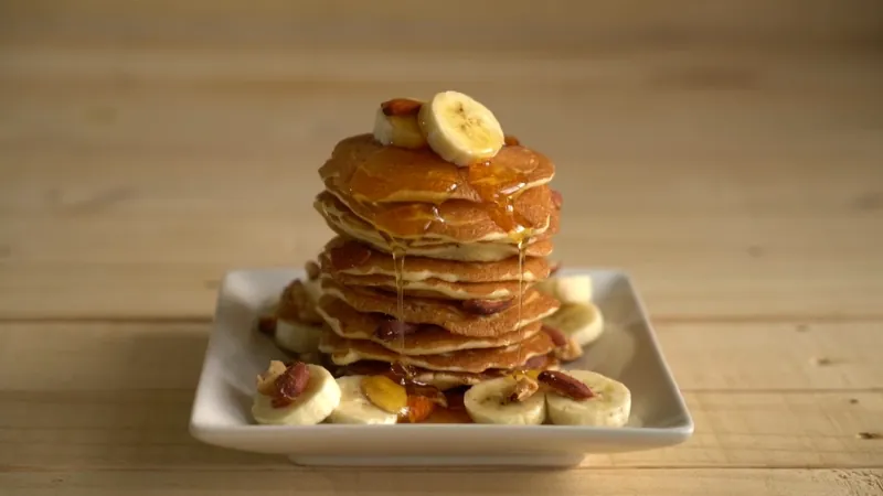 Stack of banana pancakes with syrup, banana slices, and pecans on a plate