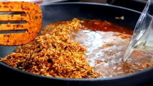 Water being poured into a pan of toasted, spiced rice using a measuring cup, with a wooden spatula resting on the side