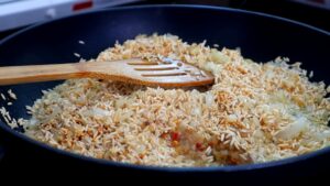 Rice, chopped onions, and spices being sautéed in oil with a wooden spatula in a black pan