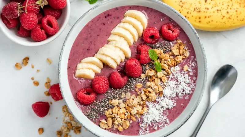 An acai bowl is topped with banana slices, raspberries, granola, chia seeds, and shredded coconut