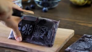 A person cuts open a frozen acai packet on a wooden cutting board with a knife