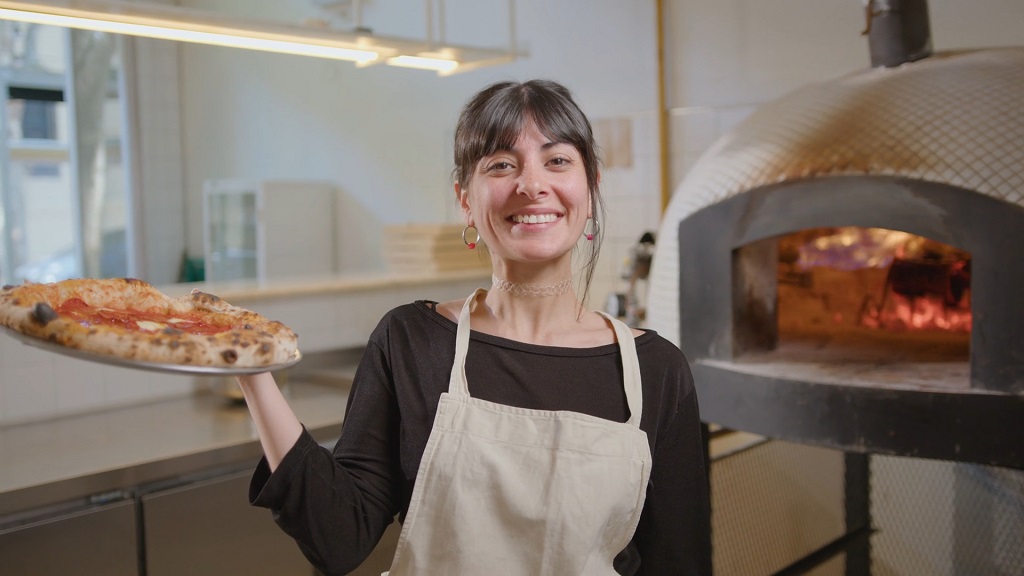 Smiling baker holding fresh pizza in front of oven