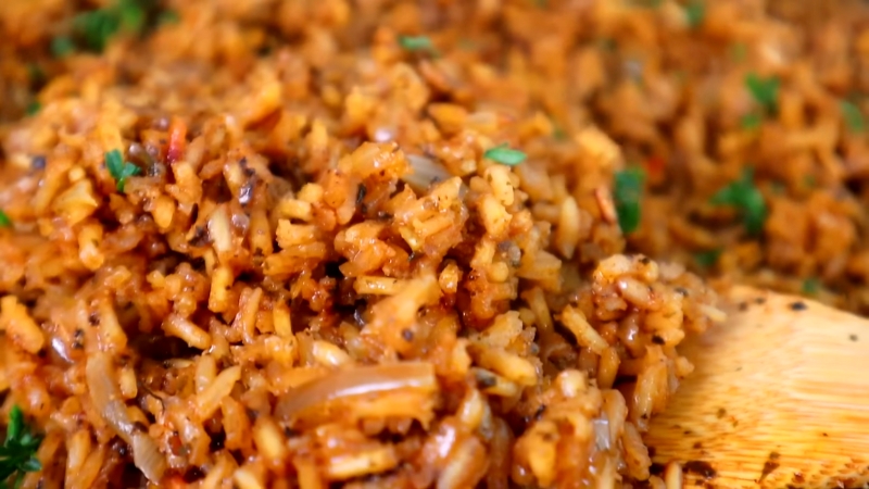 Close-up of seasoned rice mixed with onions, herbs, and spices, being stirred with a wooden spoon