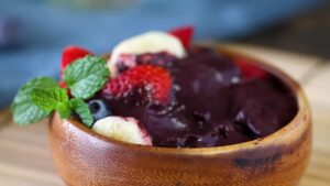A finished acai bowl is topped with sliced bananas, strawberries, blueberries, and fresh mint in a wooden bowl