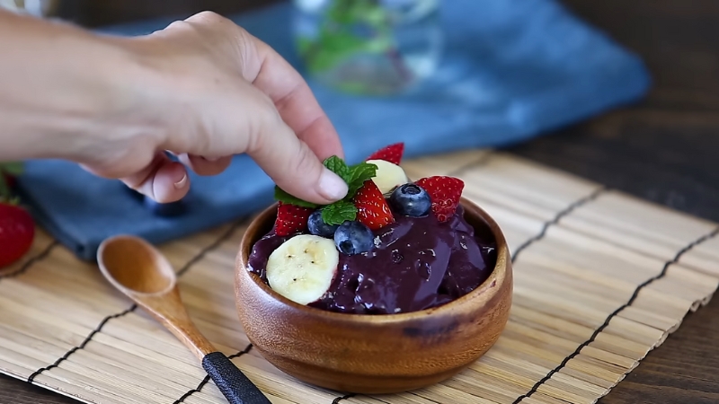 A hand places mint leaves on top of an acai bowl garnished with banana slices, strawberries, and blueberries