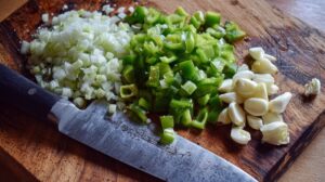 Chopped white onion, diced green bell pepper, and peeled garlic cloves on a wooden cutting board with a chef's knife