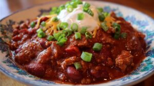 A hearty bowl of thick chili garnished with shredded cheddar cheese, sour cream, and chopped green onions, served in a floral-patterned dish