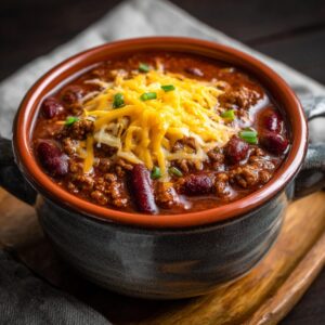 A steaming bowl of thick chili with ground beef, kidney beans, shredded cheddar cheese, and green onions served in a rustic ceramic bowl
