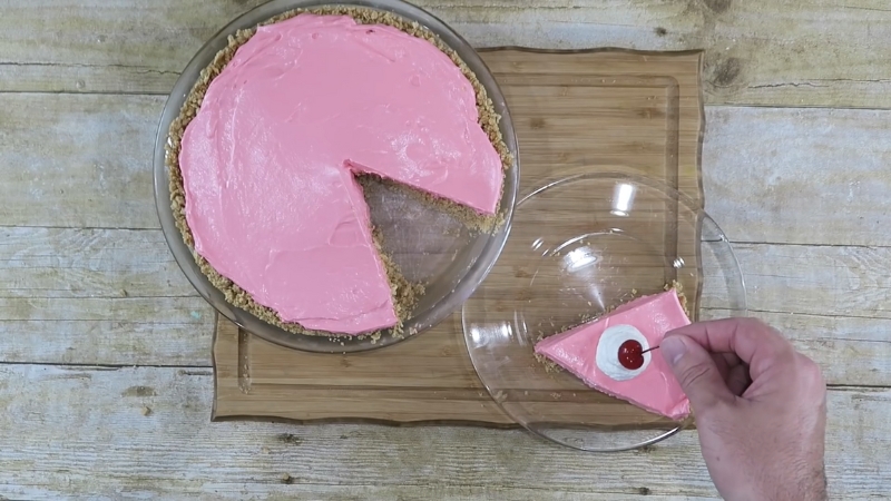 A slice of pink No-Bake Kool-Aid Pie is placed on a glass plate, topped with whipped cream and a cherry