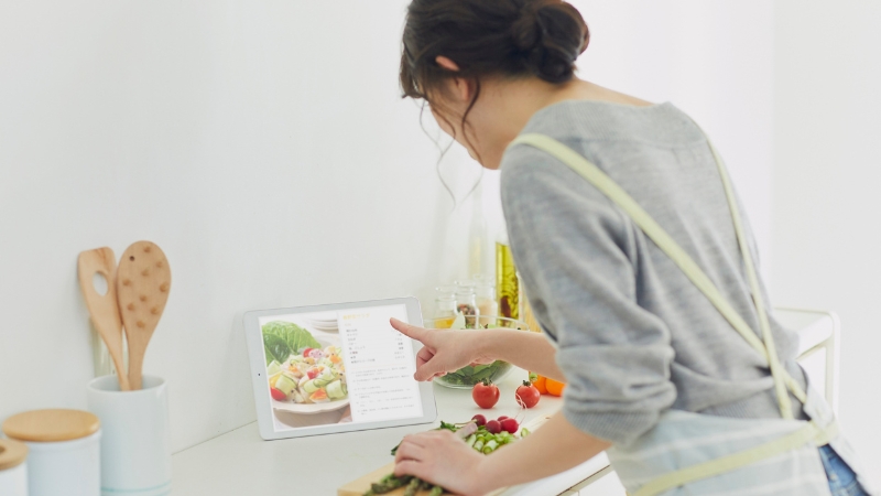 A woman in an apron follows a recipe on a tablet while preparing fresh vegetables in her kitchen