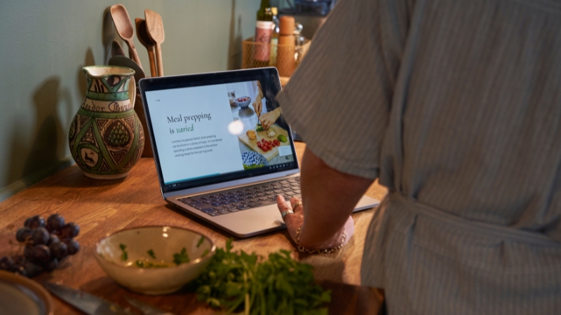 A person follows a recipe on a laptop while preparing fresh ingredients on a wooden kitchen counter